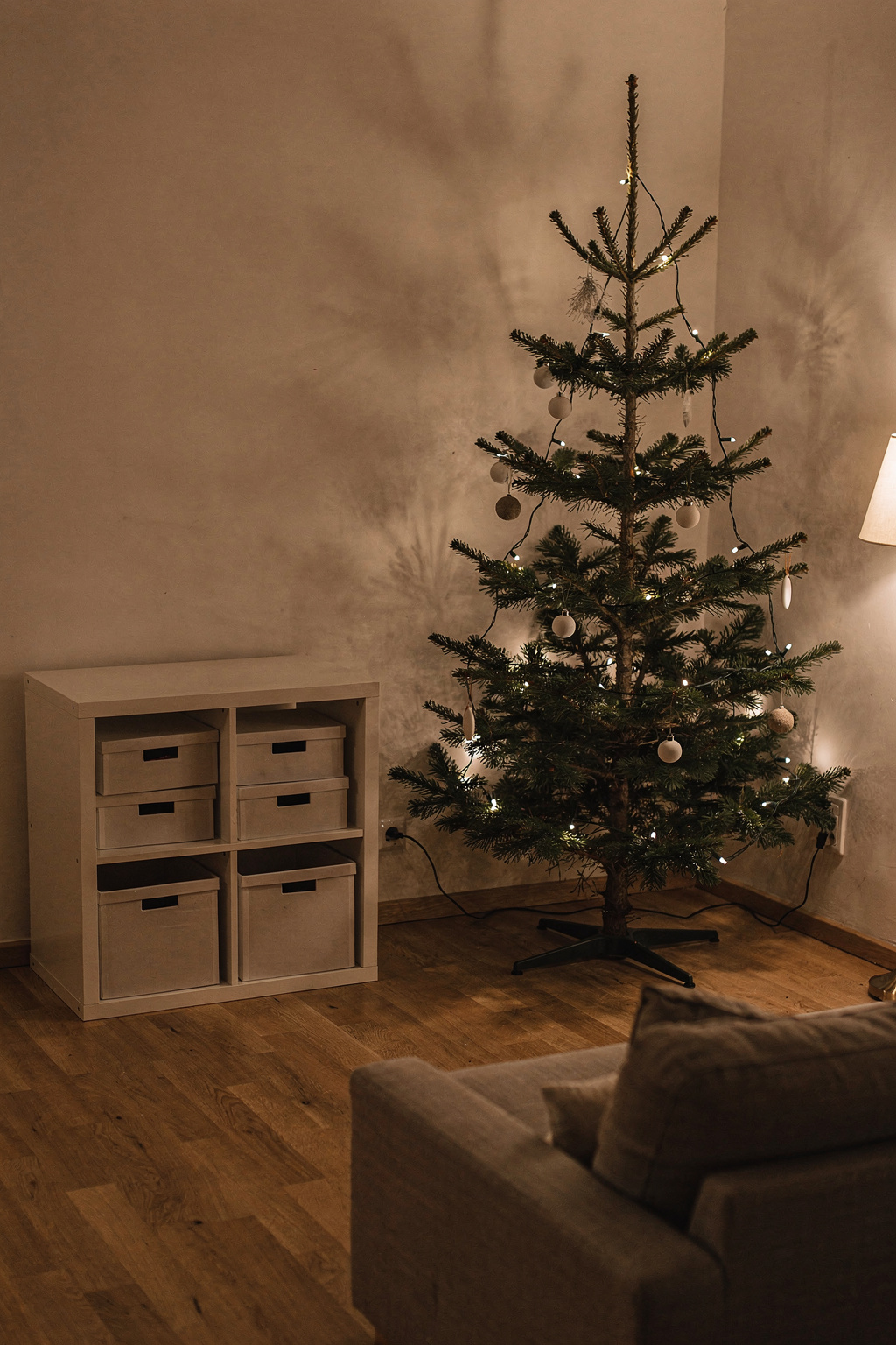 Ein schlanker Weihnachtsbaum steht in der Ecke eines kleinen Wohnzimmers mit wenig Mobiliar.