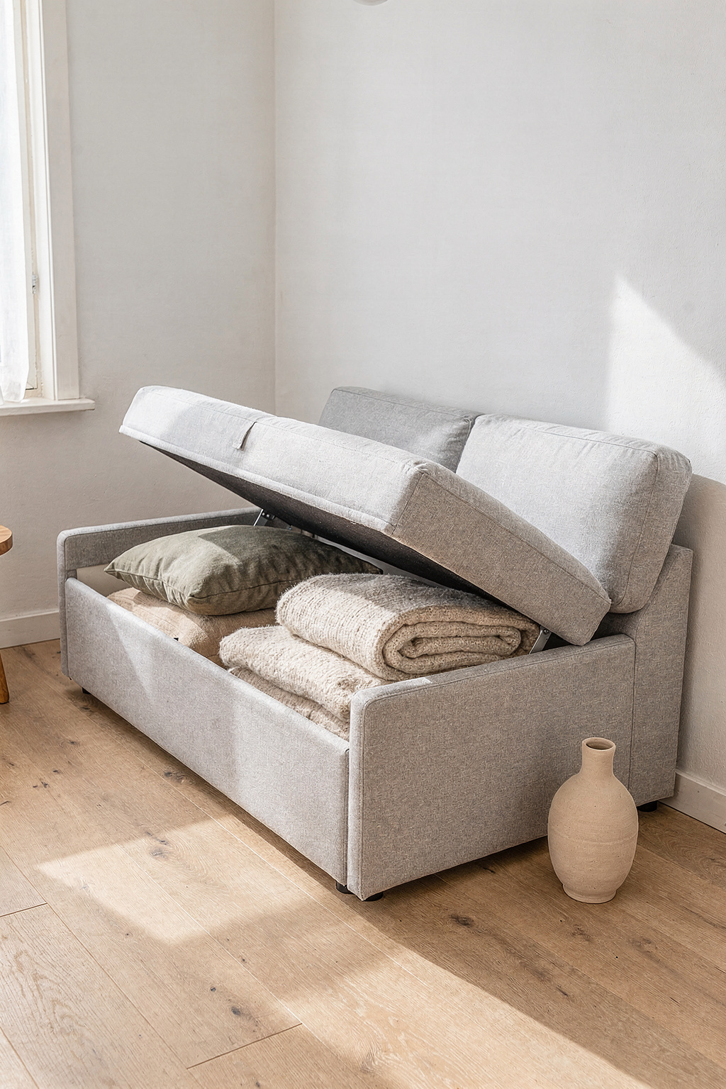 Ein hellgraues Stauraum Sofa mit geöffnetem Deckel zeigt gefaltete Decken im Inneren, Skandinavisches Wohnzimmer.