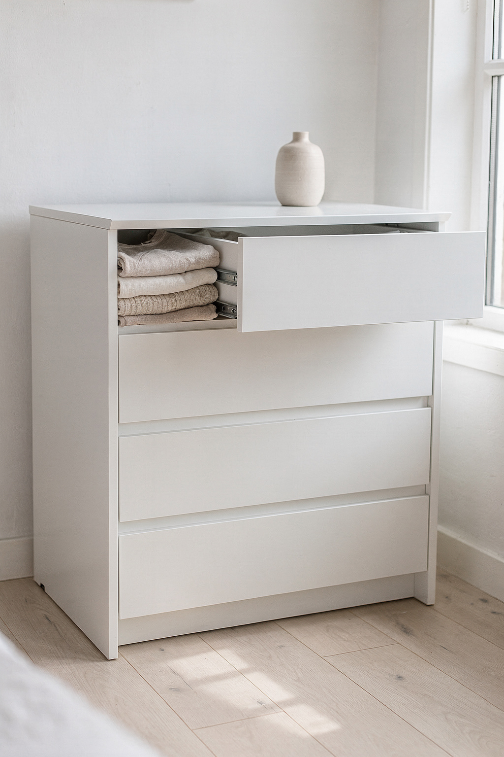 Eine weiße Kommode mit vielen Schubladen steht in einem hellen Schlafzimmer mit Holzboden.